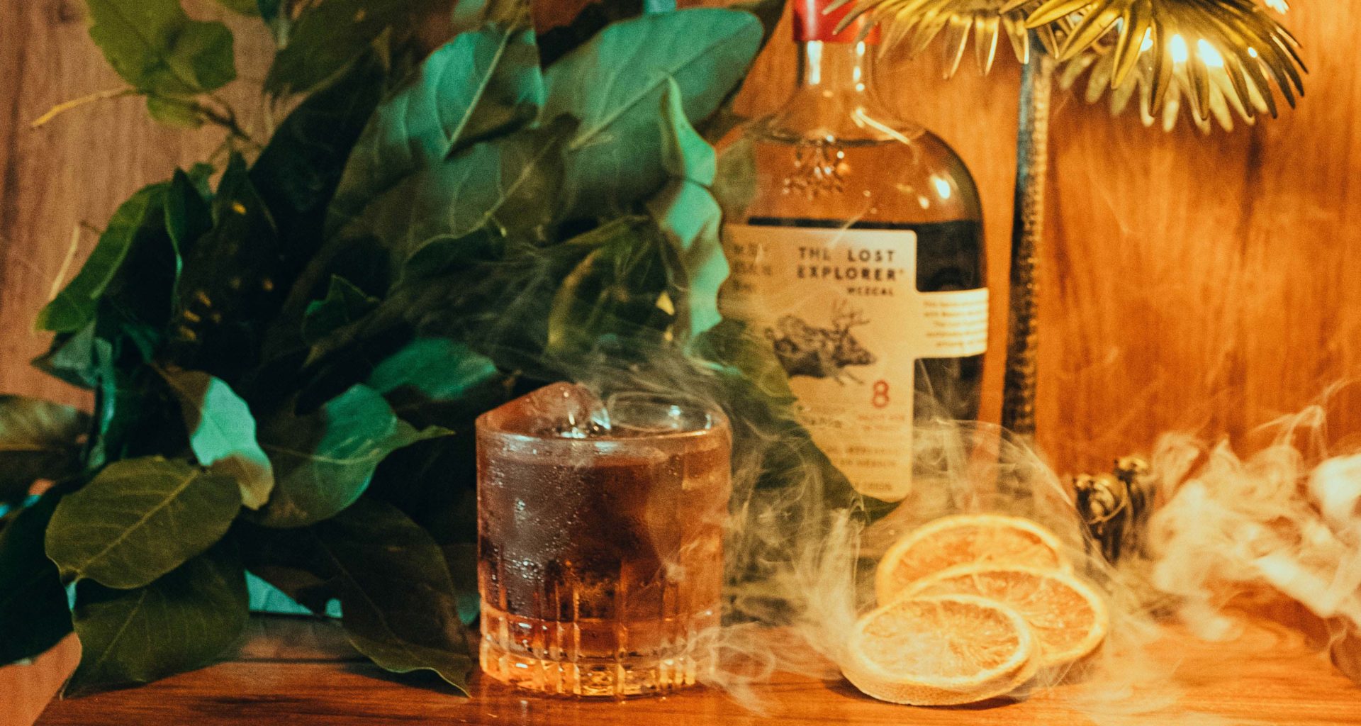 Cocktail with smoke and dehydrated orange slices on one side with a background of plants and a bottle of The Lost Explorer Espadín